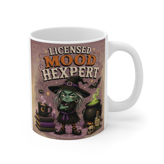 Licensed Mood Hexpert funny witch 11oz ceramic coffee mug featuring a cute green witch, bubbling cauldron, bats, and Halloween fantasy artwork.